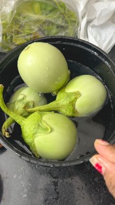 some green fruit in a black bowl on a table with other food items and a woman's hand