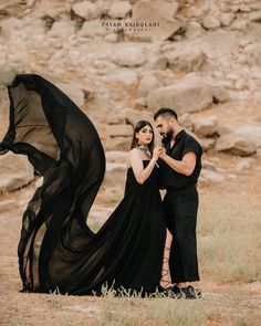 a man and woman standing next to each other in front of a rock wall with a black dress on