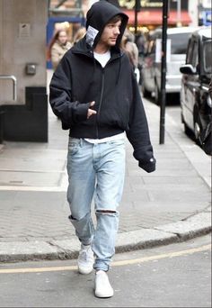 a man walking down the street wearing ripped jeans and a black hoodie with his hands in his pockets