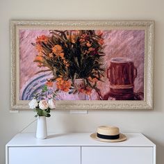 a painting hanging on the wall next to a vase with flowers in it and a hat on top