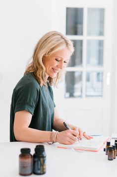 a woman sitting at a table writing on a notebook with bottles around her and an open book in front of her