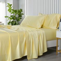 a bed with yellow sheets and pillows in a room next to a plant on a table