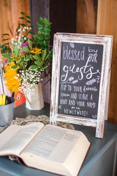 an open book sitting on top of a table next to a vase with flowers and a chalkboard sign