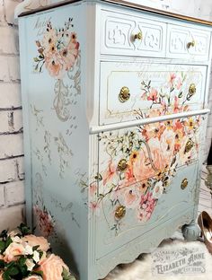 an ornate painted dresser with flowers on the drawers and gold trimmings, sits next to a white brick wall