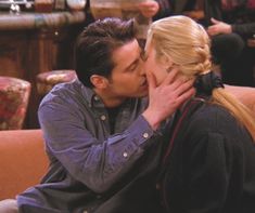 a man and woman kissing each other while sitting on a couch with people in the background