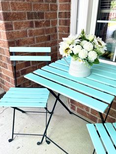 a blue table and two chairs with white flowers in a vase on the ground next to a brick wall