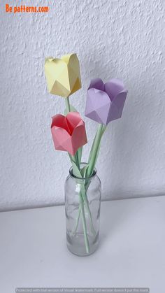 three origami flowers in a glass vase on a table with white wall background