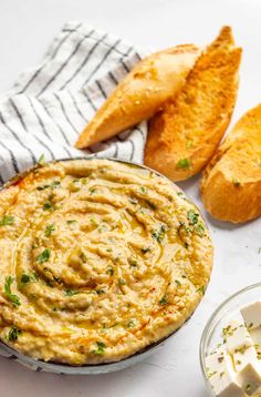 a bowl of hummus next to bread and butter on a white tablecloth with two slices of bread in the background