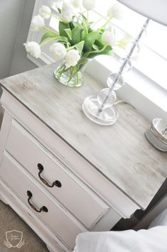 a white dresser with flowers in a glass vase on it's top and an open window behind it