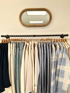 clothes hanging in front of a mirror on a rack with wooden hangers and an oval mirror above it