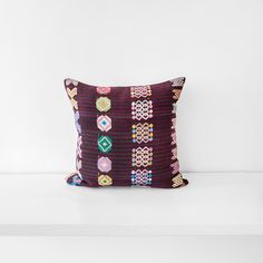 a purple pillow with multicolored designs on it sitting on a white shelf next to a vase