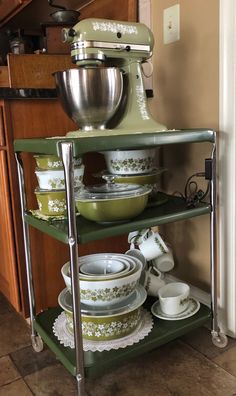 a green mixer sitting on top of a metal shelf filled with bowls and plates next to a wooden cabinet