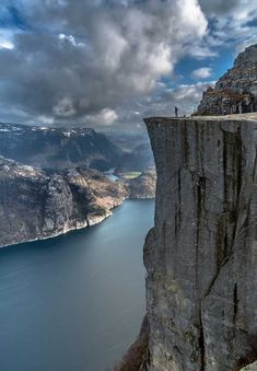 a man standing on top of a cliff next to a body of water under a cloudy sky