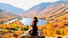 a woman sitting on top of a rock next to a valley filled with autumn colored trees