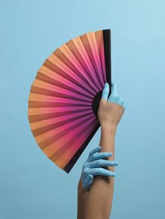 a woman's hand with blue painted hands holding an orange and pink fan over her face