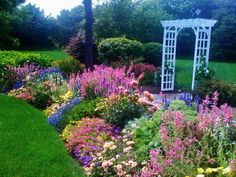 a garden filled with lots of flowers next to a white pergolated arbor on top of a lush green field