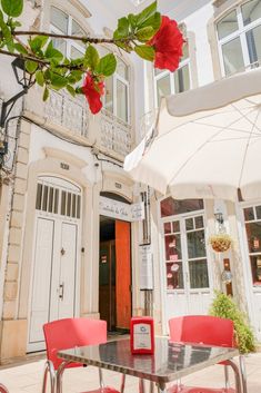 two red chairs sitting under an umbrella in front of a building with white doors and windows