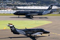 two air new zealand airplanes on the tarmac at an airport, with buildings in the background