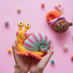 a hand holding a small crocheted snail on top of a pink surface next to other items