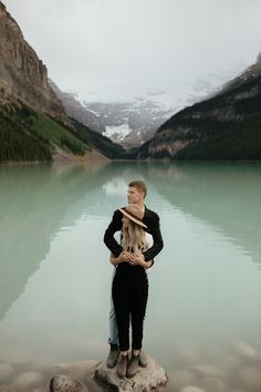 a man and woman standing next to each other on rocks in front of a lake