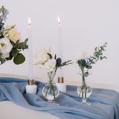 three vases filled with flowers on top of a blue table cloth next to two candles