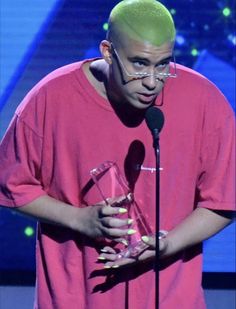 a man with green hair holding an award in front of a microphone and wearing a pink shirt