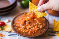 a hand holding a tortilla chip over a bowl of salsa with chips on the side