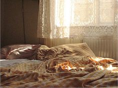 an unmade bed in front of a window with sunlight coming through the curtains on it