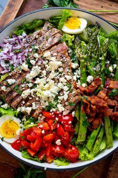 a salad with steak, asparagus, tomatoes, eggs and bacon in a bowl