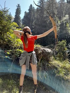 a woman in red shirt and grey skirt holding an arrow over her head while standing next to trees