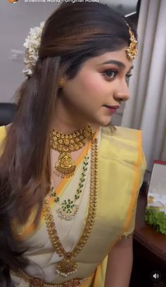 a woman wearing a yellow and white sari with gold jewelry on her neck, in front of a window