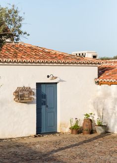 a small white building with a blue door and window on the outside, next to potted plants