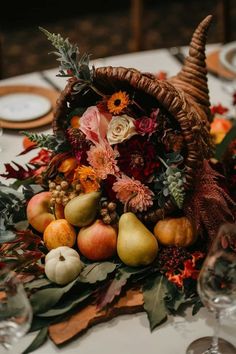 an arrangement of fruit and flowers in a basket on a table with place settings for dinner