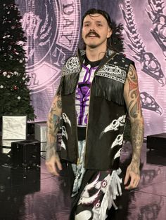 a man with tattoos standing in front of a christmas tree wearing a black leather vest