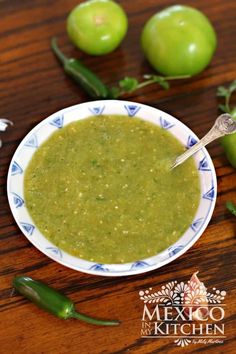 a bowl filled with green salsa sitting on top of a wooden table next to peppers