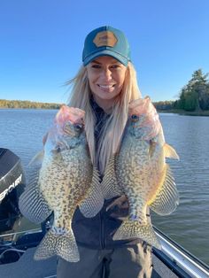 a woman holding two large fish in her hands while standing on a boat at the water's edge