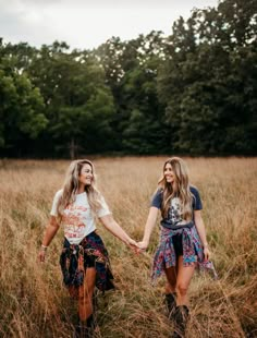 two young women holding hands and walking through tall grass in the middle of a field