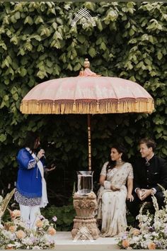 two people are sitting under an umbrella in front of flowers and greenery on the ground