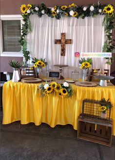 a yellow table with sunflowers and greenery on it, in front of a cross