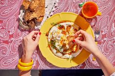 two people eating food from a yellow plate on a pink tablecloth with orange cups and utensils