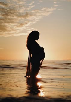 a pregnant woman standing in the ocean at sunset with her arms around her waist and head