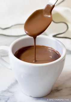 a spoon full of chocolate sauce being poured into a white cup with the liquid in it