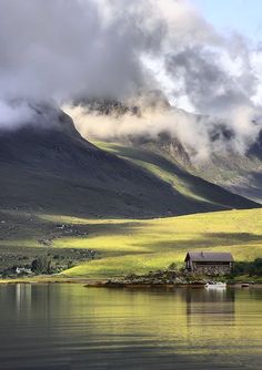 a house on the shore of a lake with mountains in the background and clouds hovering over it