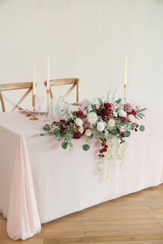 an arrangement of flowers and greenery on a white table cloth with two chairs in the background