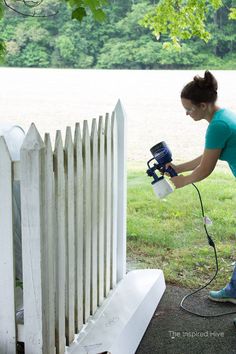 a woman using a paint sprayer on a white picket fence in front of a lake