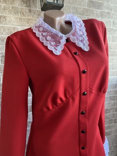 a woman's red jacket with white lace collar and buttons on the front, standing in front of a brick wall