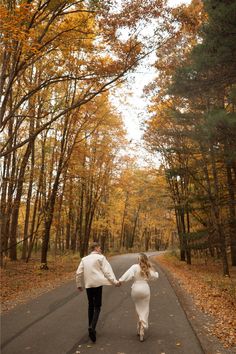 a man and woman holding hands while walking down a road surrounded by trees in the fall