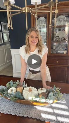 a woman standing in front of a wooden table with white pumpkins and greenery