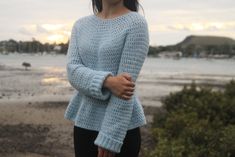 a woman standing in front of the ocean with her hand on her hip, wearing a blue knitted sweater