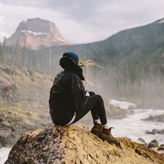 a woman sitting on top of a rock next to a river and mountains in the background